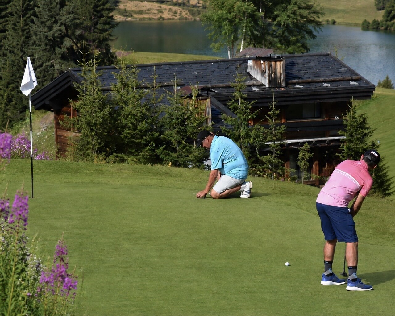 Golfers taking part in a competition on the green of the Courchevel Golf Club, with an alpine chalet, mountain lake and alpine flowers in the background.
