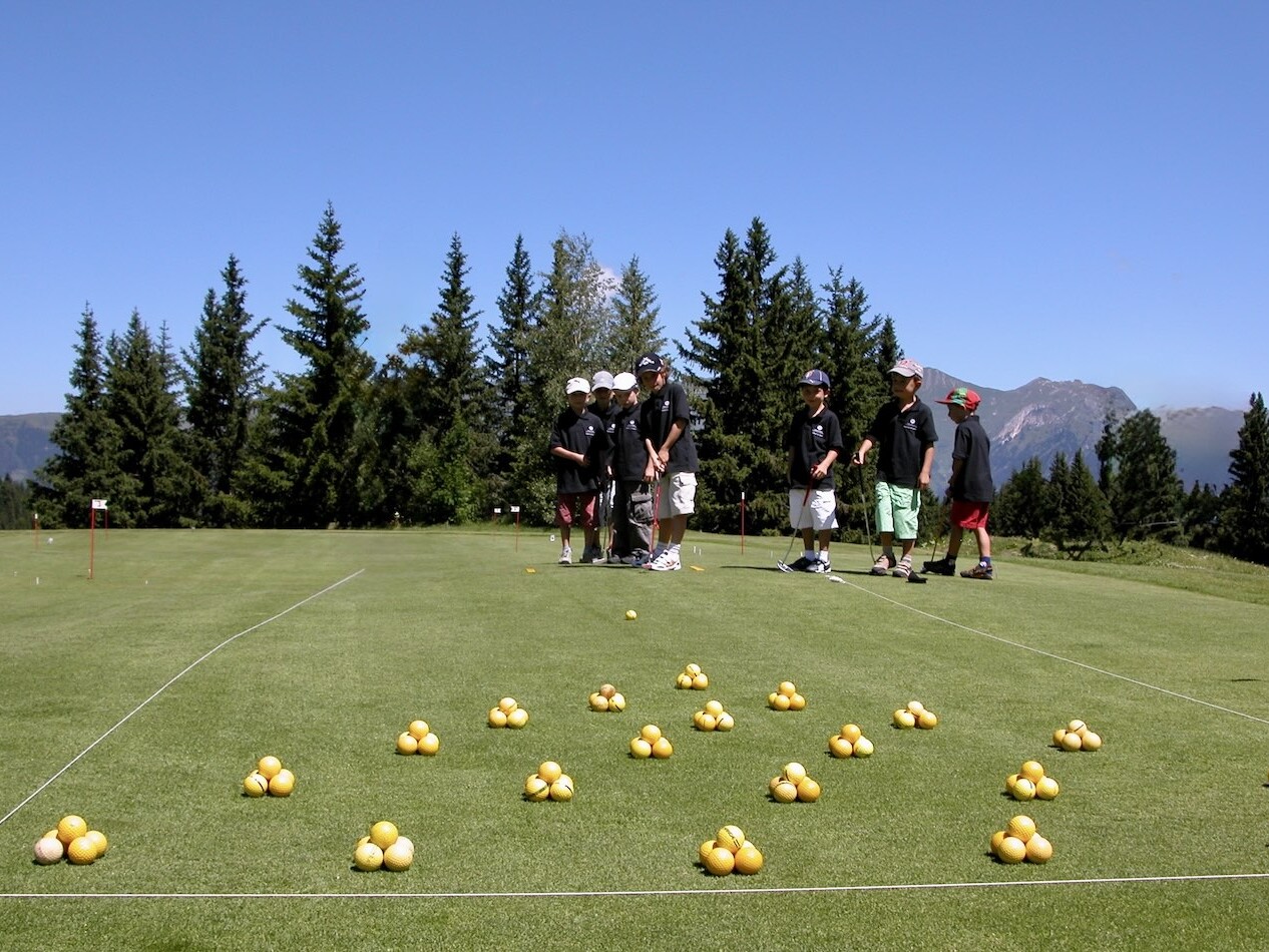 Enfants participant à un stage junior de golf sur le putting green du Golf Club de Courchevel avec les montagnes des Alpes en arrière-plan.